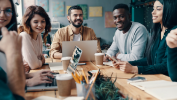 A diverse, smiling team of Impimpi Tech professionals collaborating during a meeting around a wooden office table.
